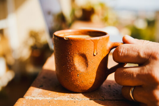 Hand holding a rustic clay mug with coffee on a balcony