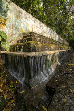 View of the tiered fountain at Parque Nacional Barranca del Cupatitzio with cascading water flowing over mossy stone steps and a mural wall in the background in Uruapan, Michoacan, Mexico.