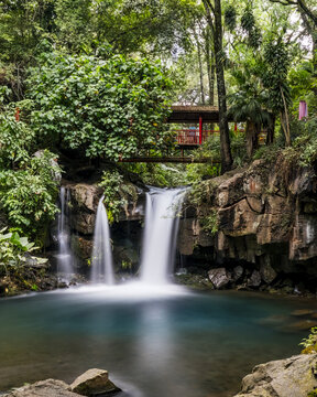 View of the waterfall at Parque Nacional Barranca del Cupatitzio with a wooden bridge, lush tropical vegetation, and a turquoise pool in Uruapan, Michoacan, Mexico.