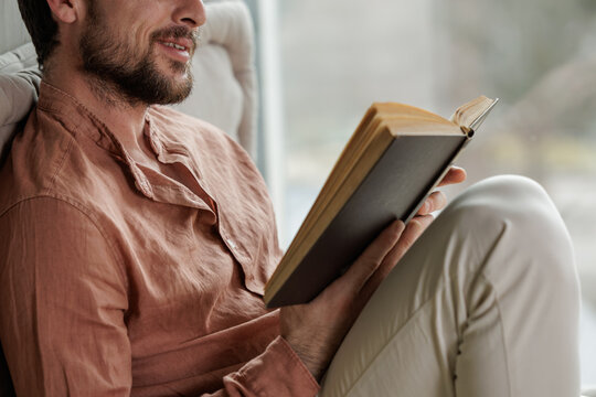 Happy young man smiling while reading a book by a window during leisure time