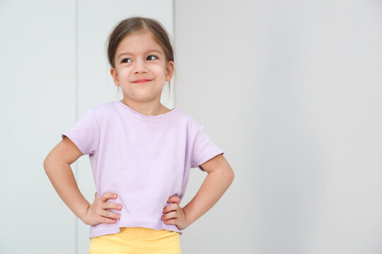 Cheerful Young Girl Poses With Hands On Hips In Front Of Bright White Wall, Wearing Lavender Shirt And Yellow Pants, Showing Confident Playful Mood, Clean Studio Lighting, Simple Composition