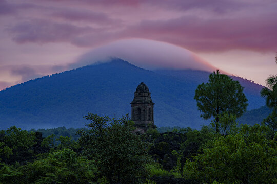 View of the historic church tower of San Juan Parangaricutiro rising above lush trees with the Paricutin volcano and a lenticular cloud under a purple sunset sky in Angahuan, Michoacan, Mexico.