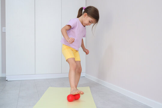 Focused Young Girl Balancing Barefoot On Yoga Mat While Standing On Red Spiky Massage Ball In Bright Home Interior, Wearing Purple Shirt And Yellow Shorts For Fitness, Coordination