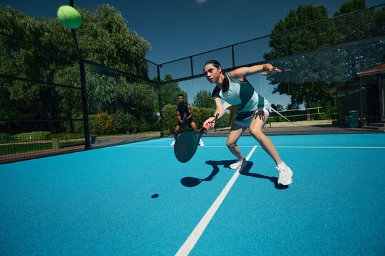 Female padel player in stylish blue sportswear reaching for ball during outdoor game. Concept of athletic apparel marketing, functional fashion, summer sport lifestyle.
