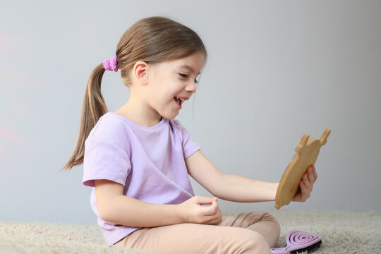 Smiling young girl sitting on soft rug and looking at wooden mirror, enjoying quiet indoor play, side profile, bright natural light, simple home background, cheerful childhood moment
