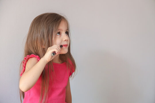 Young Girl In Pink Shirt Applies Lip Makeup With Small Cosmetic Brush While Looking Upward In Side Profile Against Plain Light Background, Showing Focused Expression, Long Hair, Soft Natural