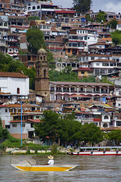 View of a fisherman in a traditional butterfly net boat on Lake Patzcuaro with the hillside town of Janitzio featuring dense houses and a church tower in Janitzio, Michoacan, Mexico.