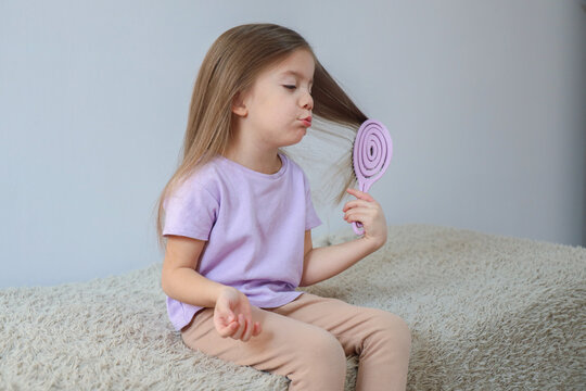 Young girl sitting on soft bed brushing long hair with lilac detangling brush, wearing pastel clothing, calm home grooming moment with gentle expression and cozy minimal interior