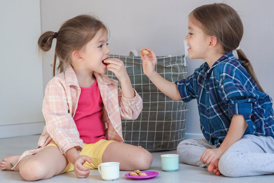 Two Young Girls Sit On Floor Indoors, Sharing Small Snacks And Drinks While Smiling At Each Other During Relaxed Home Playtime, Showing Friendship, Togetherness, Casual Clothing