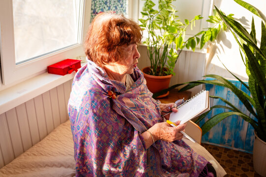 Elderly retired Caucasian woman makes handwritten notes in a notebook in a cozy living room. Productivity, learning, and managing daily tasks concept. Side view