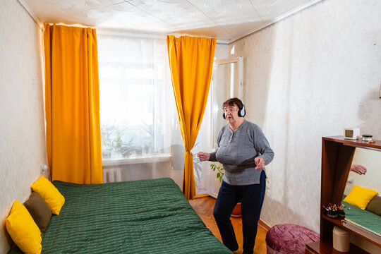 Senior Caucasian woman smiles while dancing to music in headphones inside a peaceful room. Active leisure inspires positive emotions and enjoyable retirement. Wide shot