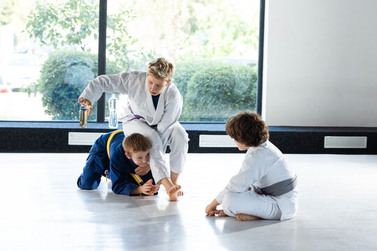 Blonde judo coach helps two children complete controlled ground exercise. Concept of beginner grappling lessons, safe practice guidance and youth clubs.