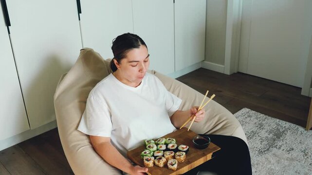 Adult woman rests in beige lounge chair, eating sushi from wooden tray with chopsticks inside clean modern home interior, with relaxed posture, soft natural light, and quiet domestic mood
