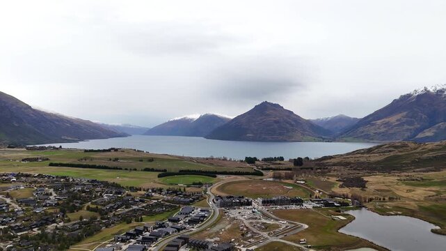 Aerial drone footage sliding left across Lake Wakatipu and the Jack&rsquo;s Point landscape, revealing rolling farmland, wetlands, and dramatic mountain ranges beneath overcast skies.