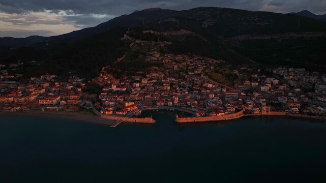 Drone aerial shot approaching the historic Venetian port and medieval town of Nafpaktos illuminated by warm golden sunrise light, with castle hill and calm sea, Gulf of Corinth, Greece.