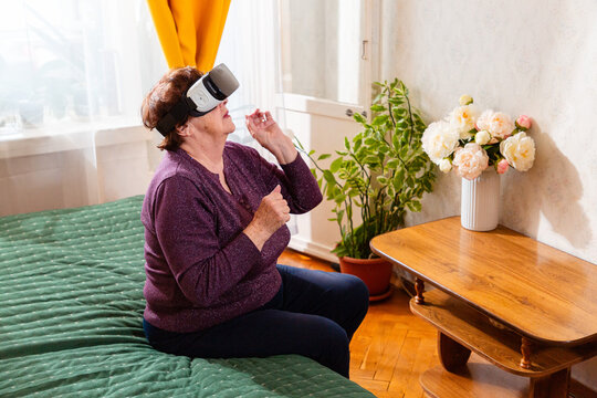 Caucasian senior woman enjoying VR entertainment while sitting comfortably on a bed. Modern technology provides an engaging pastime. Side view