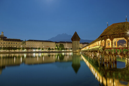View of the historic Chapel Bridge and Wasserturm illuminated at dusk, reflecting in the calm waters of the Reuss River with the distant mountains in Lucerne, Lucerne, Switzerland.