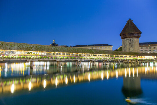View of the illuminated Chapel Bridge and Wasserturm reflecting in the Reuss river at night in Lucerne, Switzerland.