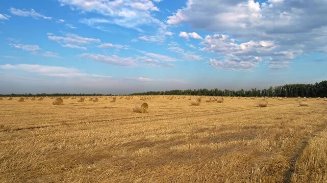 Drone follows parallel farm tracks toward a single round straw bale growing larger in the foreground while distant rolls a dark tree border and layered clouds define the harvested plain