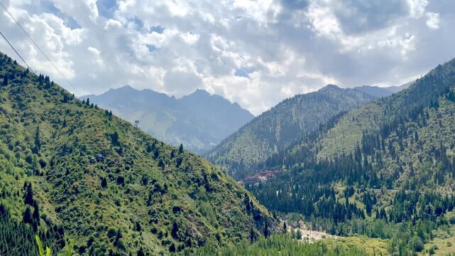 4K panoramic view of lush green mountains near Almaty, Kazakhstan, seen from Kok Tobe viewpoint. Scenic mountain valley, forested slopes and dramatic cloudy sky create a beautiful summer landscape.