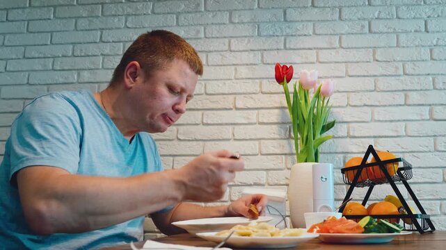 Adult man in light blue shirt eating at home table beside white brick wall, tulips, fruit basket, plated vegetables, and soft indoor light in calm side profile kitchen setting