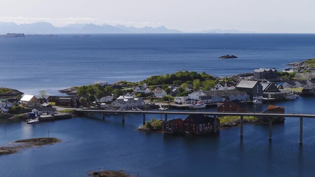 Aerial view shows a long concrete bridge spanning across the sea to connect small coastal islands and fishing town buildings in Lofoten, Norway on a sunny day.