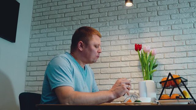 Adult man seated beside dining table indoors, eating quietly with cutlery near plate, beside fruit basket, tulips, white brick wall, warm light, and calm contemporary home decor