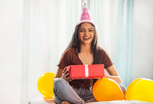 Portrait of a cheerful young woman wearing birthday hat and holding birthday gift in hand