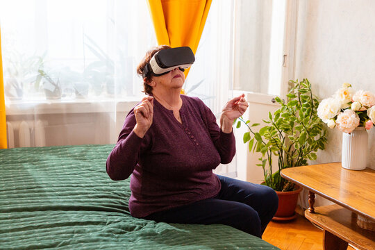 Senior retired woman wearing VR glasses enjoys realistic virtual adventures while sitting on a sofa. Home recreation and technology for older generations.