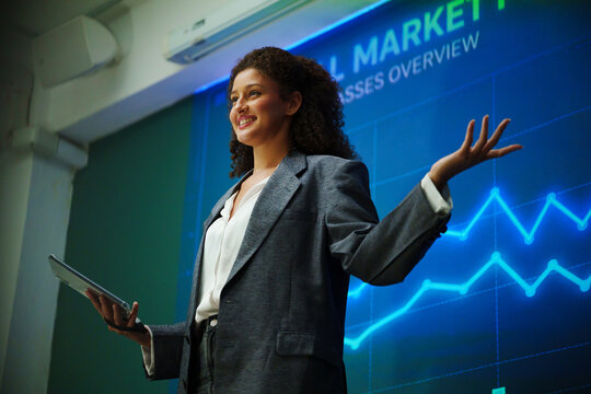 Confident businesswoman presenting financial market charts with a tablet during a corporate seminar, speaking to an audience about investment analysis, performance, strategy and growth trends.