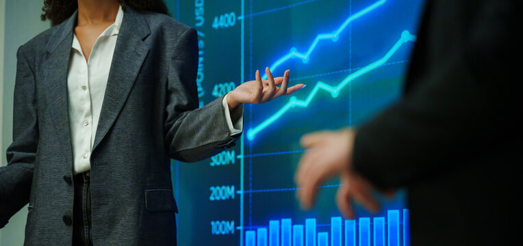 Businesswoman presenting financial charts and rising market trends on a digital screen during a corporate seminar, explaining investment performance, analytics, strategy and economic growth.
