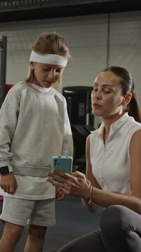 Vertical video of Caucasian female trainer showing fitness app on smartphone to Caucasian child in gym. Footage is suitable for promoting family wellness programs and healthy lifestyle apps