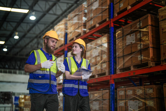 Male warehouse supervisor with a beard and his female coworker in protective workwear hold a two way radio while checking the racks in a large logistics distribution centre with tall.