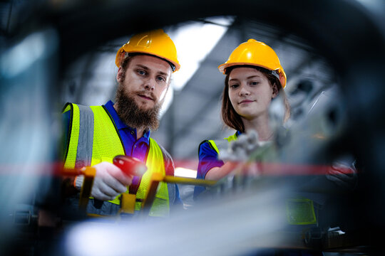Two warehouse employees, a bearded man and a young woman, wearing yellow hard hats point a barcode reader at components between machine frames in an industrial plant that stores and.