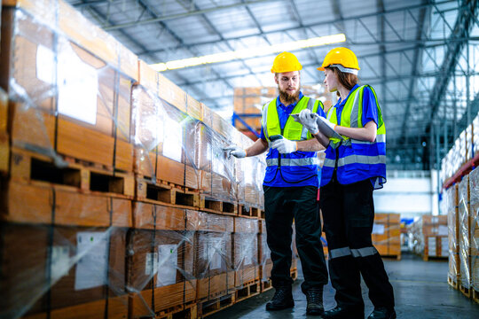 Male warehouse supervisor with a beard and his female coworker in protective workwear review pick lists in the middle of the storage aisle in a large fulfilment centre with rows of stacked.