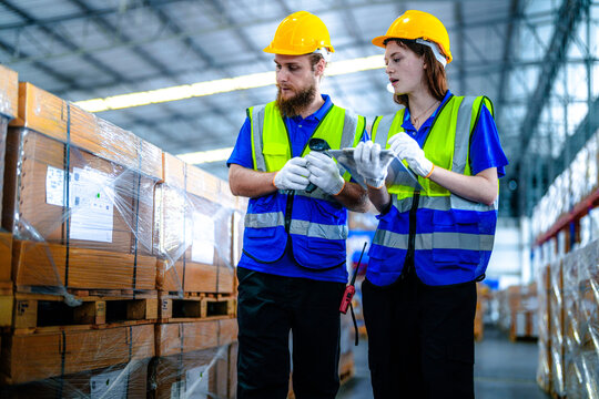 Male warehouse supervisor with a beard and his female coworker in protective workwear aim a glowing barcode reader at stacked cartons in a spacious logistics centre filled with shrink.