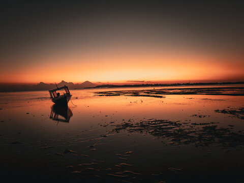 View of a small boat in the shallow waters of Gili Trawangan at sunset, with a distant volcano silhouette against an orange sky in Pemenang, West Nusa Tenggara, Indonesia.