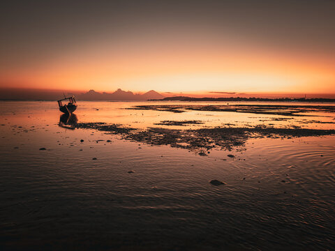 View of a small boat in the calm shallow waters of Gili Trawangan at sunset with a mountain silhouette under a warm golden sky in Pemenang, West Nusa Tenggara, Indonesia.