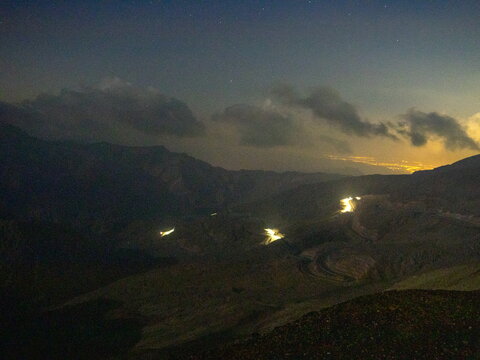 View of the winding road on Jebel Jais illuminated by car headlights at night under a starry sky in Ras Al Khaimah, United Arab Emirates.