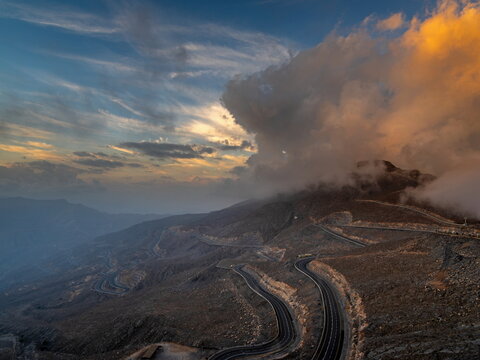 View of the winding mountain road of Jebel Jais at sunset with dramatic clouds over the rocky landscape in Ras Al Khaimah, United Arab Emirates.