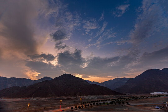 View of the rugged Hajar Mountains at sunset with a dramatic cloudy sky and a road stretching through the desert landscape in Ras Al-Khaimah, Ras Al Khaimah, United Arab Emirates.