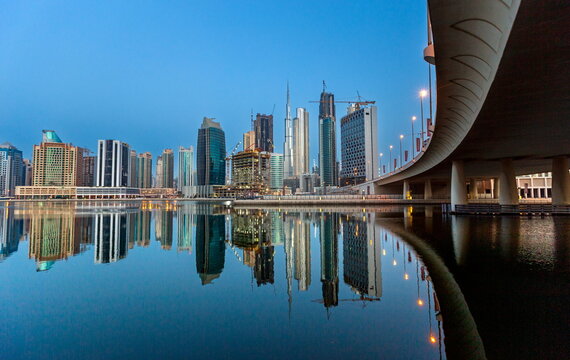 View of the Dubai skyline featuring the Burj Khalifa and modern skyscrapers reflected in the calm water at dusk with a bridge in the foreground in Dubai, Dubai, United Arab Emirates.