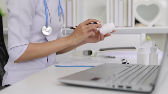 Female doctor writing prescription and examining medicine bottle. Professional female doctor with a stethoscope writing a prescription while examining a medicine bottle at her desk.