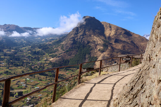 View of the Pisac archaeological site featuring a dirt path with a wooden railing overlooking the terraced agricultural valley and Andean mountains in Pisac, Cusco, Peru.