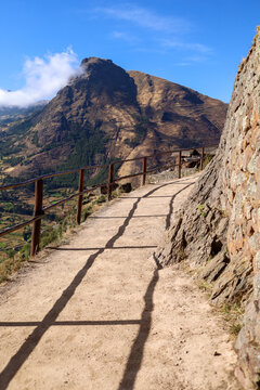 View of the ancient stone path at the Pisac archaeological site with a wooden fence and mountain views in Pisac, Cusco, Peru.