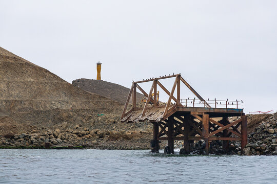 View of a weathered metal pier structure extending into the sea with birds perched on the frame, set against a coastal landscape with a yellow lighthouse in Chancay, Lima Region, Peru.