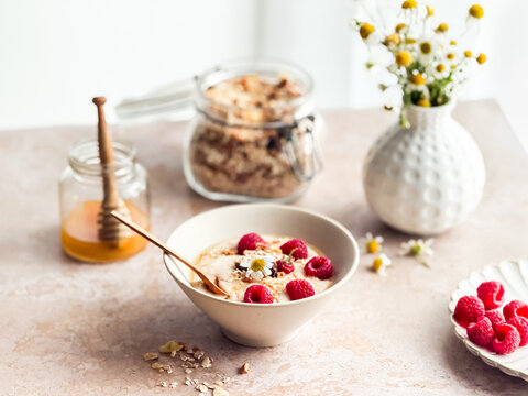 Eine Sch&uuml;ssel mit M&uuml;sli, Milch, frischen Himbeeren und Honig auf einem Tisch am Fenster. Fr&uuml;hst&uuml;ck, Zuhause.