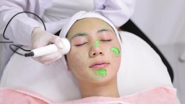 Esthetician performing ultrasonic facial treatment on a client. Relaxed woman getting a rejuvenating skin care procedure. A cosmetologist uses an ultrasonic device on her face with a green gel mask.