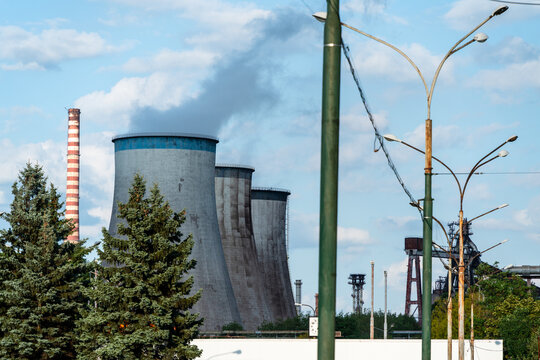 View of the industrial power plant with large cooling towers emitting smoke, framed by evergreen trees and street lamps in Dabrowa Gornicza, Silesian Voivodeship, Poland.