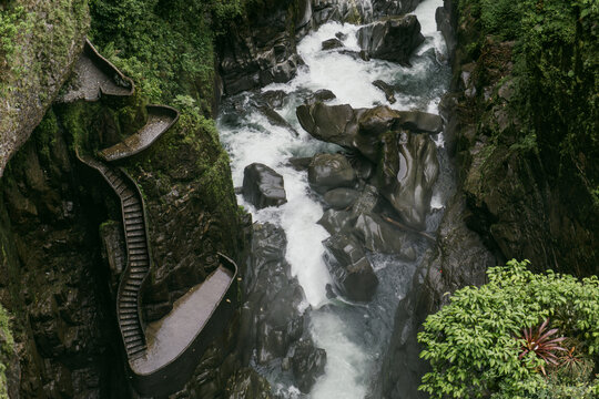 View of the stone stairs leading down to the Pailon del Diablo waterfall, surrounded by lush green vegetation and dark rock formations in Banos, Tungurahua, Ecuador.